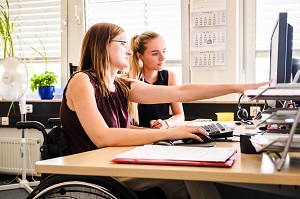 Zwei Frauen an einem Schreibtisch. Die eine, im Rollstuhl sitzend, zeigt der anderen etwas auf dem Bildschirm.