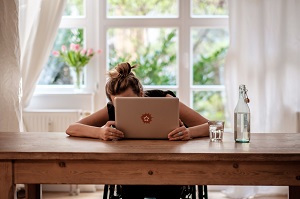 Junge Frau, im Rollstuhl sitzend, versteckt sich hiner einem Laptop.