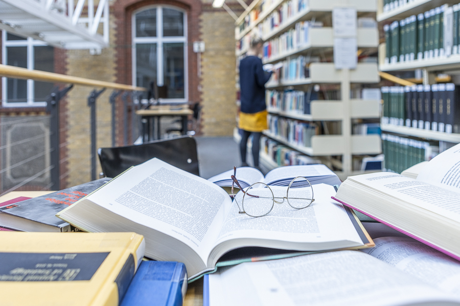 Table with many books, glasses, woman in the background, library
