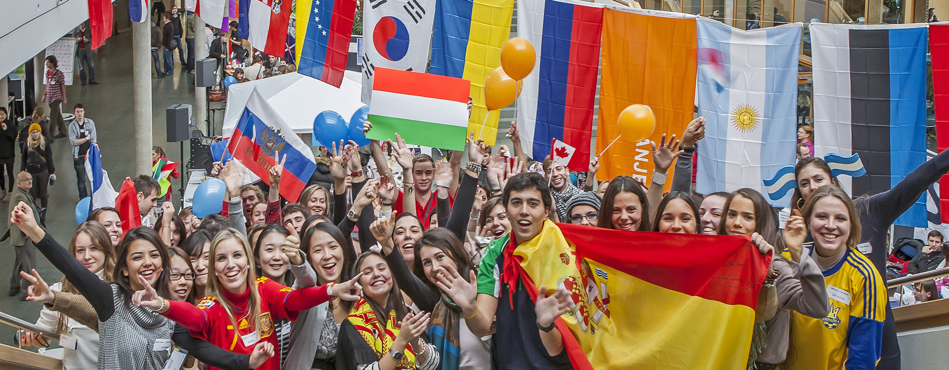 A cheerful group of young people celebrates in the Gräfin-Dönhoff-Building of the Viadrina. They are holding various flags, wearing matching clothes and waving or laughing. More flags hang in the background and colourful balloons float in the air.